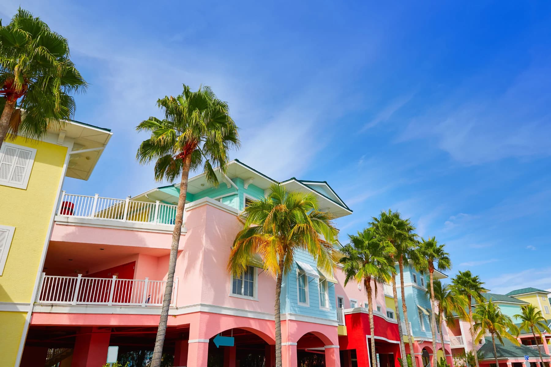 Row of colorful pastel beach buildings with balconies and palm trees lining the street under a clear blue sky.